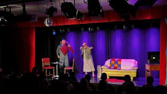 Kabarettszene im K-BUM in Liechtenstein – bunte Bühne mit Sofa, Schauspielerduo und Publikum im Saal.