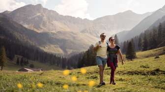 Zwei Personen wandern durch eine weite Alpwiese mit Blick auf die umliegenden Berge der Alp Valüna bei sonnigem Wetter