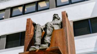 Skulptur „Hochsitz“ von Robert Indermaur: Figur sitzt erhöht auf rostfarbenem Stahlstuhl vor moderner Fassade.