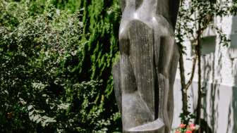Moderne Figurenskulptur aus Bronze, stehend zwischen Blumen und Sträuchern in Vaduz – Werk von Herbert Albrecht.