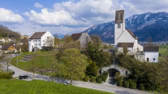 Panoramablick auf den Kirchhügel Bendern mit historischer Kirche, umgeben von gepflegten Wegen, Bäumen und traditioneller Architektur vor alpiner Bergkulisse.