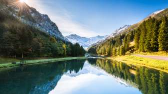 Spiegelglatter Gänglesee in Steg, umrahmt von dichten Wäldern und schneebedeckten Bergen bei Sonnenschein