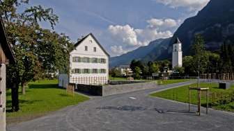 Historischer Pfarrhof mit weißer Fassade und grünen Fensterläden inmitten eines offenen Platzes, im Hintergrund die Kirche von Balzers und die umliegende Berglandschaft.
