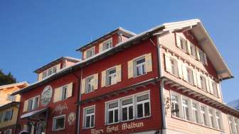 Außenansicht des Alpenhotel Vögeli Malbun mit der Elchbar im Erdgeschoss – rotes Traditionshaus mit weißer Beschriftung vor blauem Himmel.