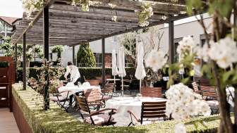 Eine elegante Terrasse mit weißen Tischen, gedeckt mit Servietten und Gläsern unter einem Holzpavillon, umgeben von üppigem Grün.