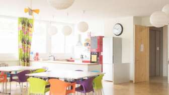 Heller Gemeinschaftsraum im müze Familienzentrum mit bunten Stühlen, runden Hängelampen und einer modernen Küche im Hintergrund.